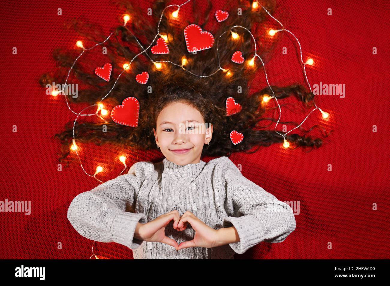 Ein Mädchen mit Girlande und roten Herzen in lockigem Haar lächelt auf rotem Hintergrund und hält ein Herz in ihren Händen. Stockfoto