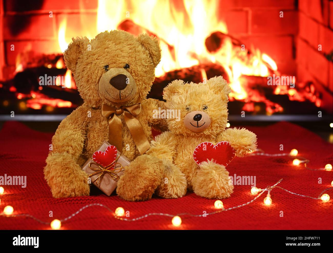 Ein Paar Teddybären sitzt umarmt vor dem Hintergrund eines roten Strickkarlots und einer Girlande aus Glühbirnen und hält zwei rote Herzen in sich Stockfoto