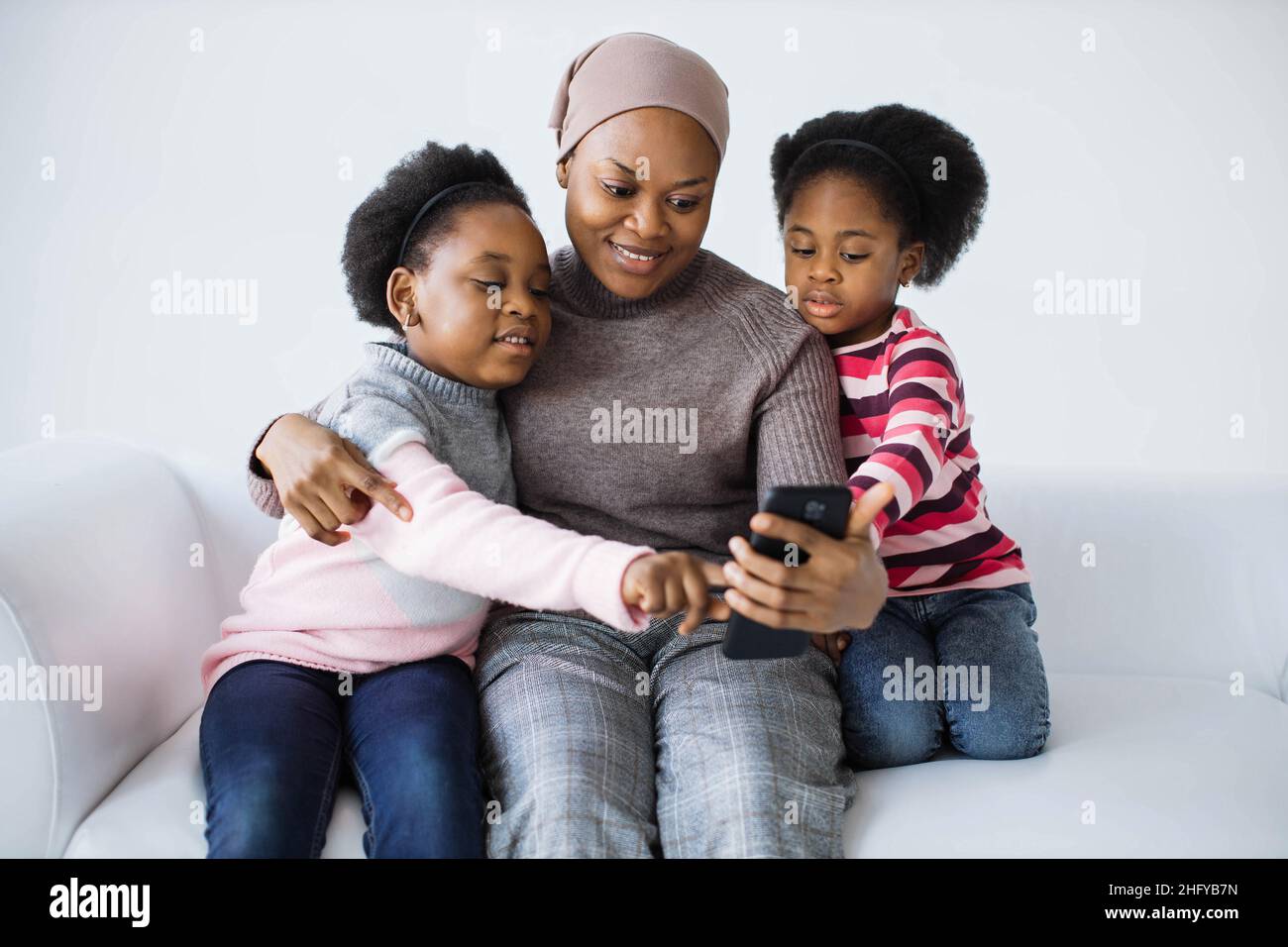 Zwei niedliche Schwestern und ihre fürsorgliche Mutter verwenden ein modernes Smartphone, um Selfie zu machen, während sie sich auf der bequemen Couch ausruhen. afroamerikanische Familie, die Spaß hat, während sie Zeit zusammen verbringt. Stockfoto