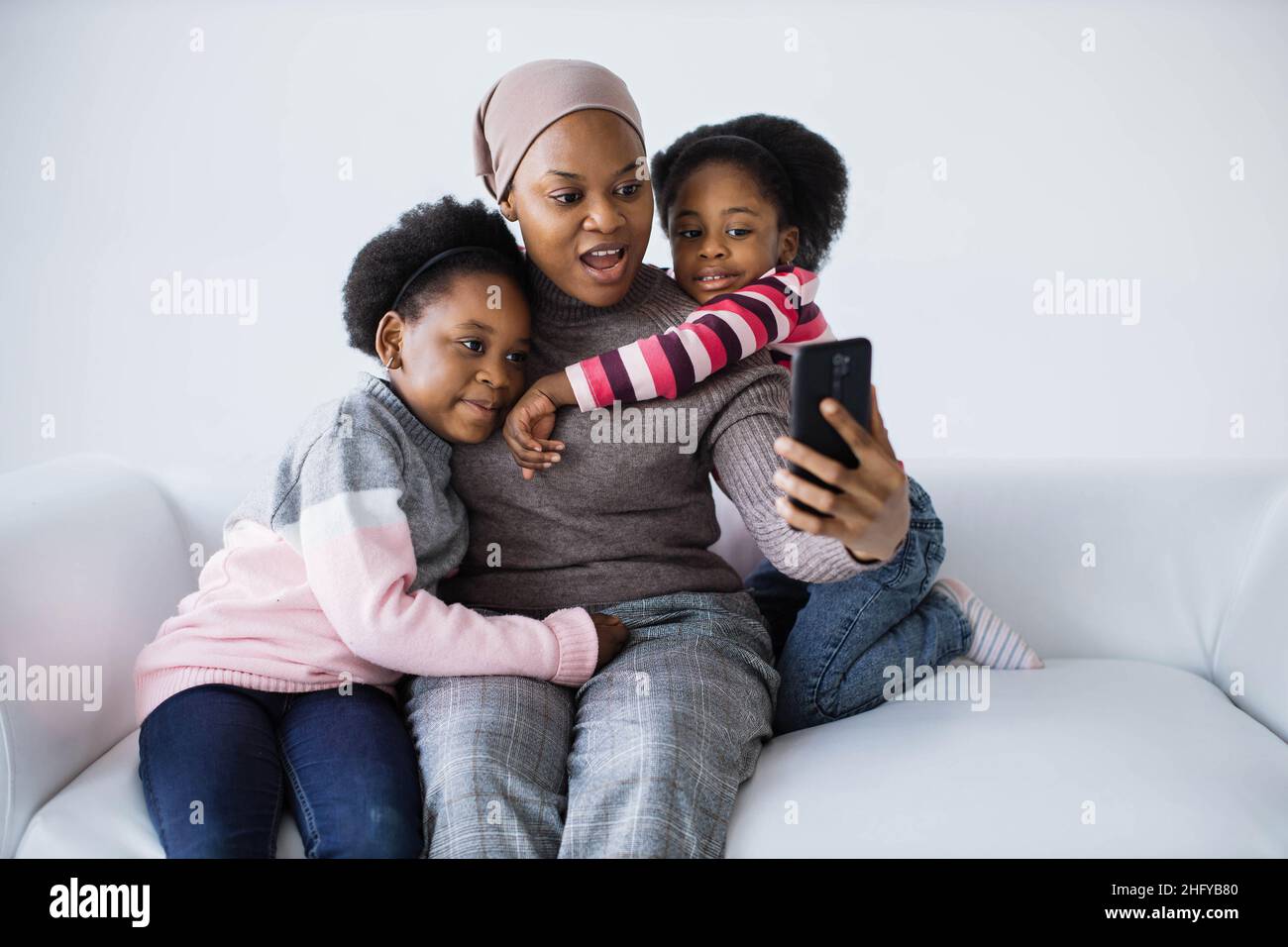afroamerikanische Frau sitzt mit zwei hübschen Tochter auf der Couch und nimmt Selfie auf dem modernen Smartphone. Weißer Studiohintergrund. Familie und Gadgets Konzept. Stockfoto