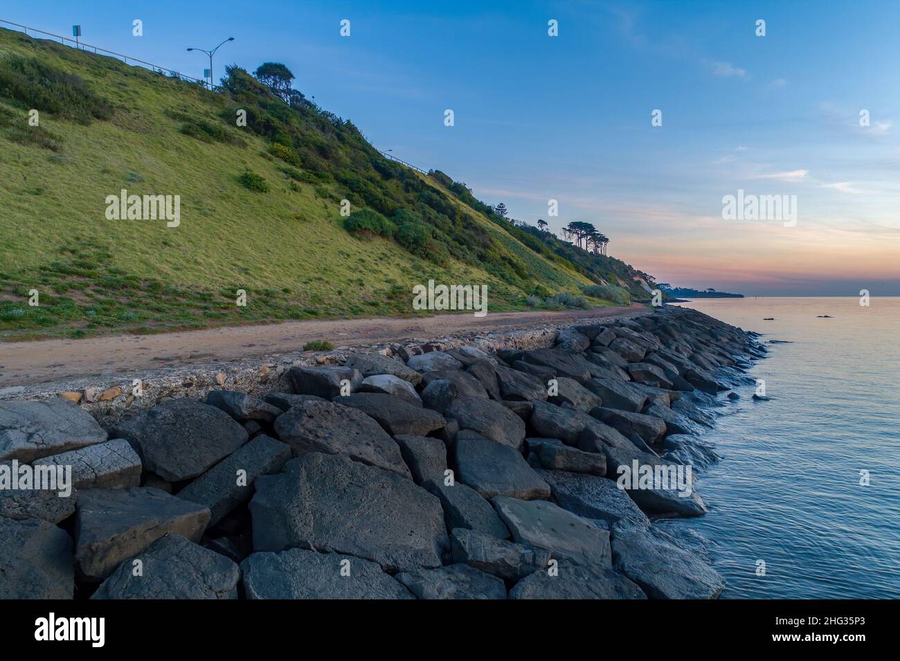 Wellenbrecher und grasbewachsener Hügel mit Bäumen bei Sonnenuntergang in Frankston, Australien Stockfoto