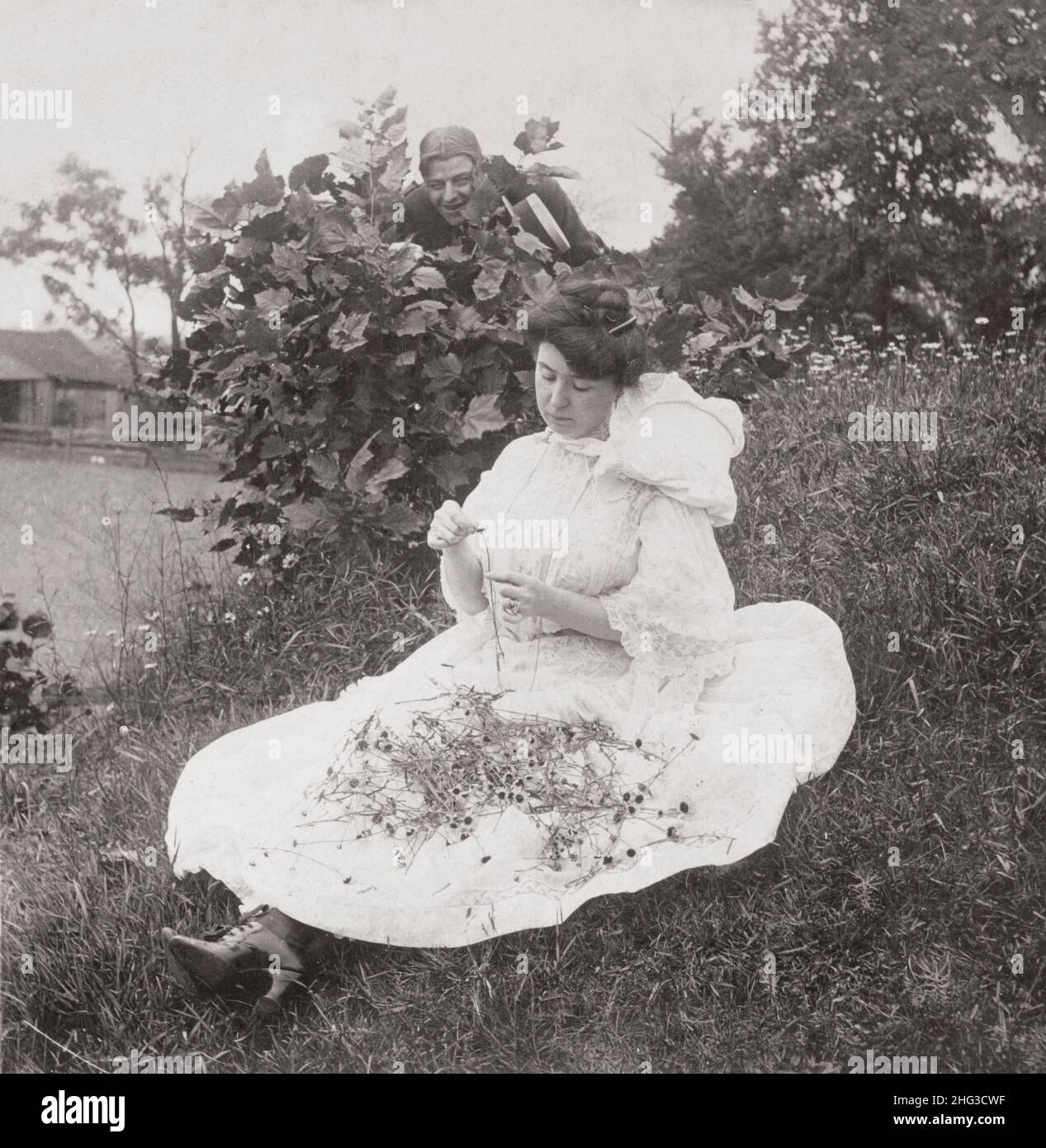 Vintage-Foto der Liebe. Er liebt mich, er liebt mich nicht. 1906 Frau saß auf dem Boden und pflückte Blütenblätter aus Blumen, während der Mann hinter dem Busch zuschaut. Stockfoto