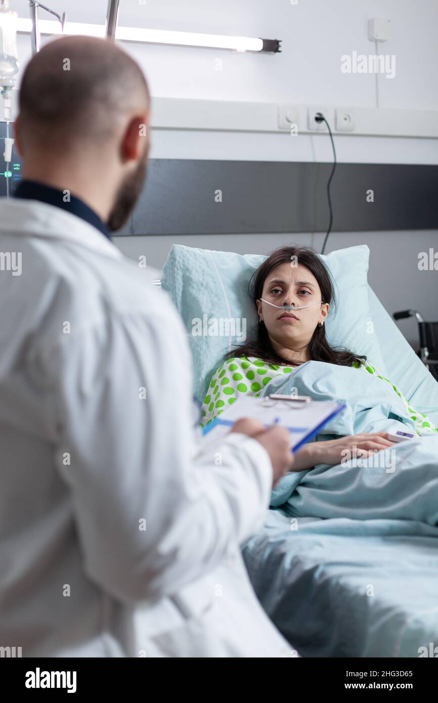 Patient mit nasaler Kanüle, der Sauerstoff auf der privaten Station erhält, sieht sich den Arzt an, der Behandlungsmöglichkeiten präsentiert, während er die Zwischenablage mit medizinischen Unterlagen hält. Frau mit niedriger sättigung von SpO2 erholt sich. Stockfoto