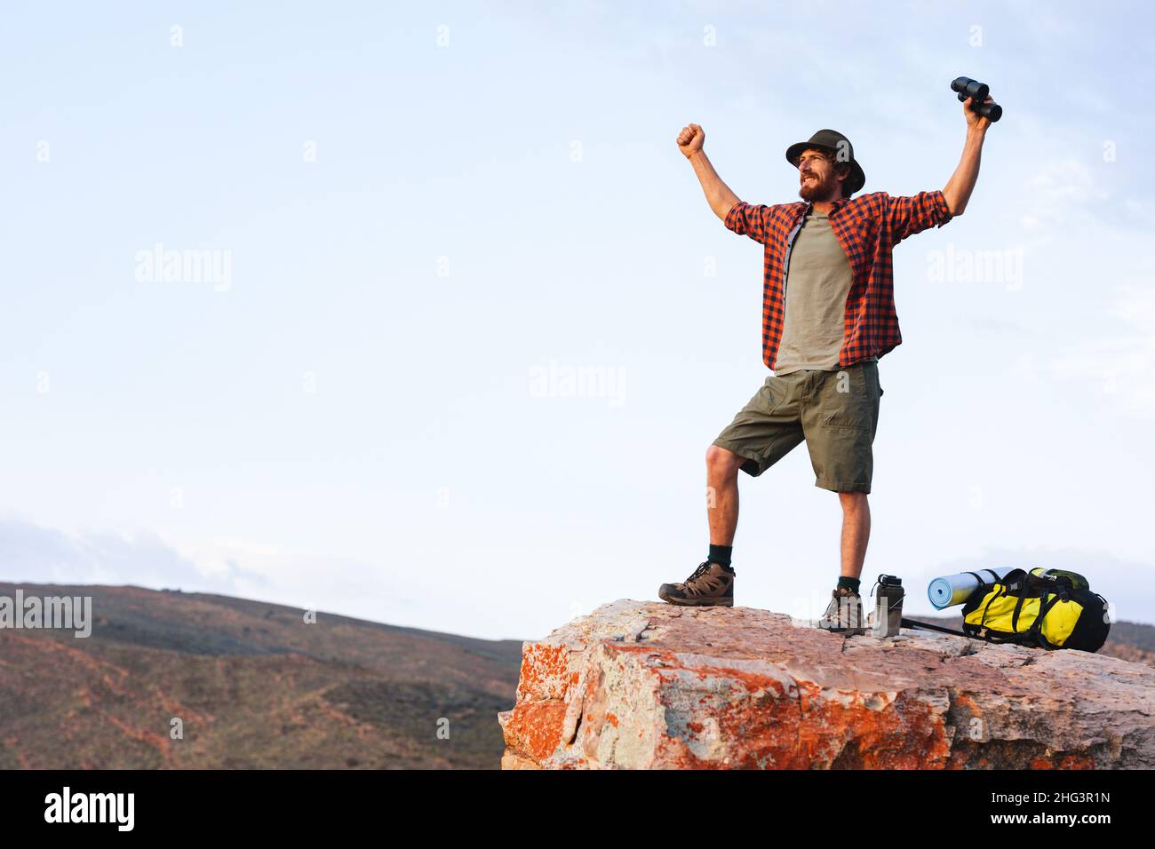 Die ganze Länge des glücklichen jungen kaukasischen Abenteurers, der bei Sonnenuntergang mit einem Fernglas auf dem Felsen jubelt Stockfoto