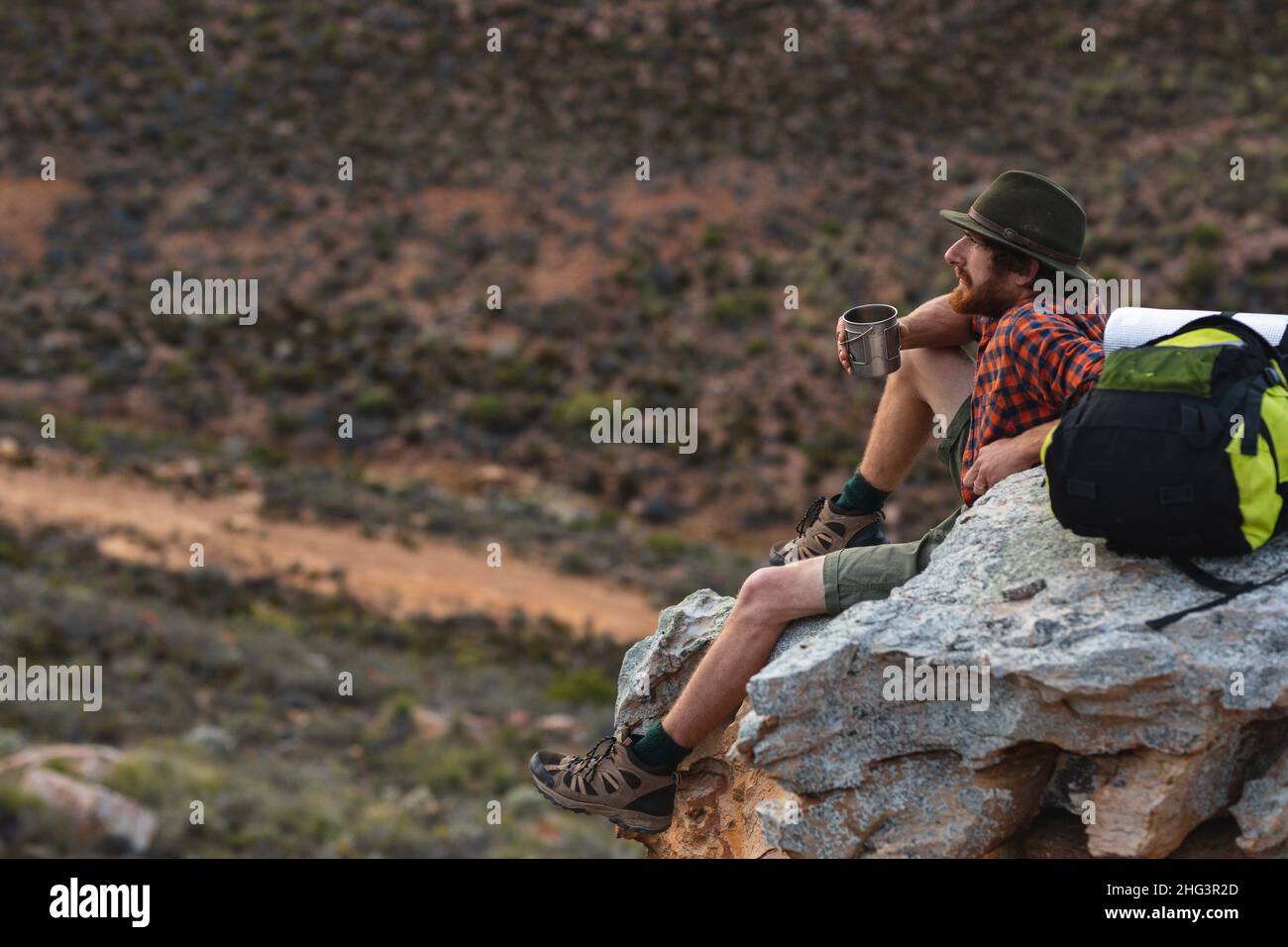 Junger kaukasischer Abenteurer, der mit einem Becher auf einer felsigen Klippe auf dem Berggipfel sitzt Stockfoto