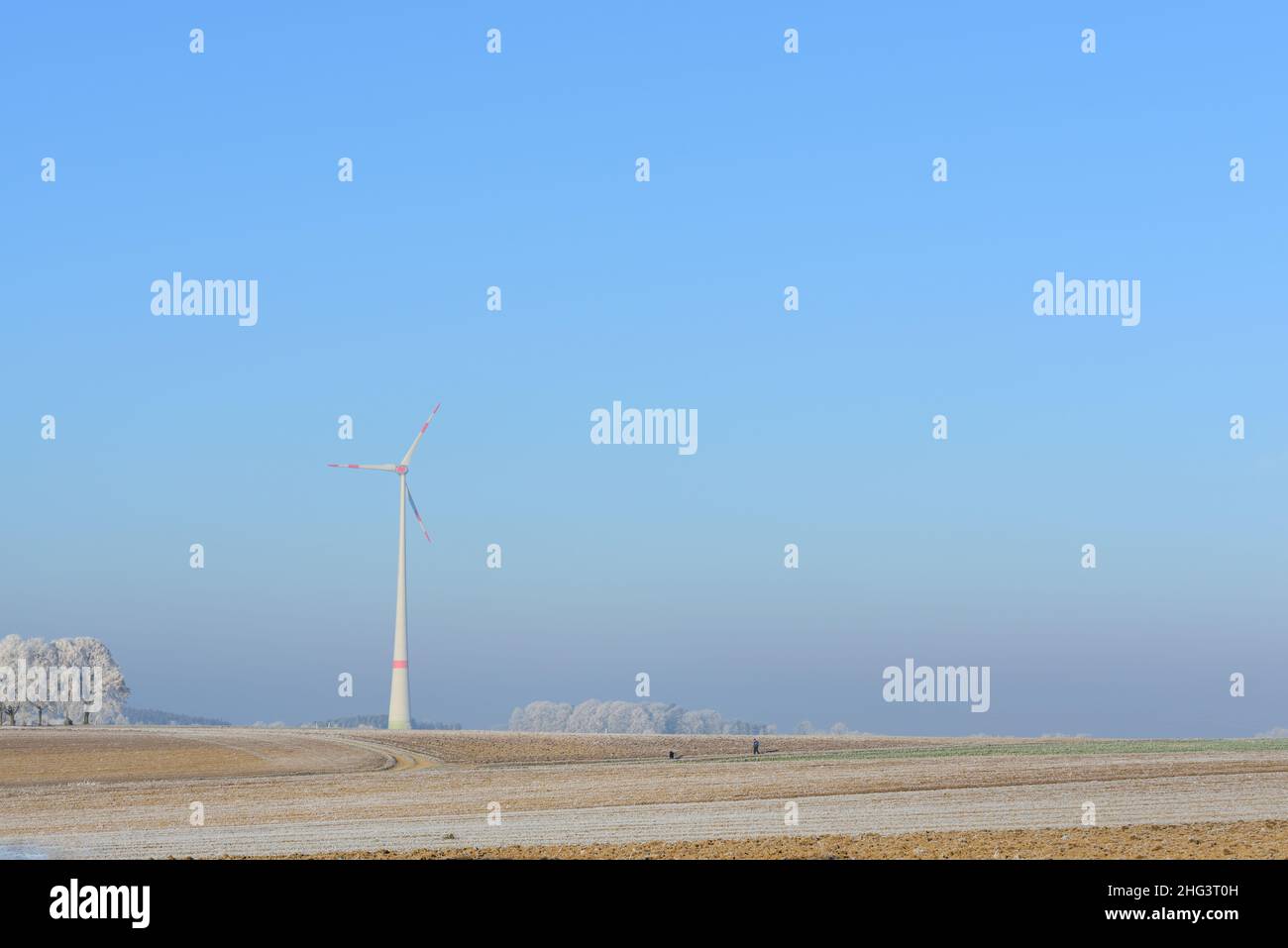 Szenische Aufnahme eines Wintertages in Bayern, mit weiten, frostbedeckten Feldern, dem blauen Himmel im Hintergrund und einer großen Windmühle zur Stromerzeugung Stockfoto