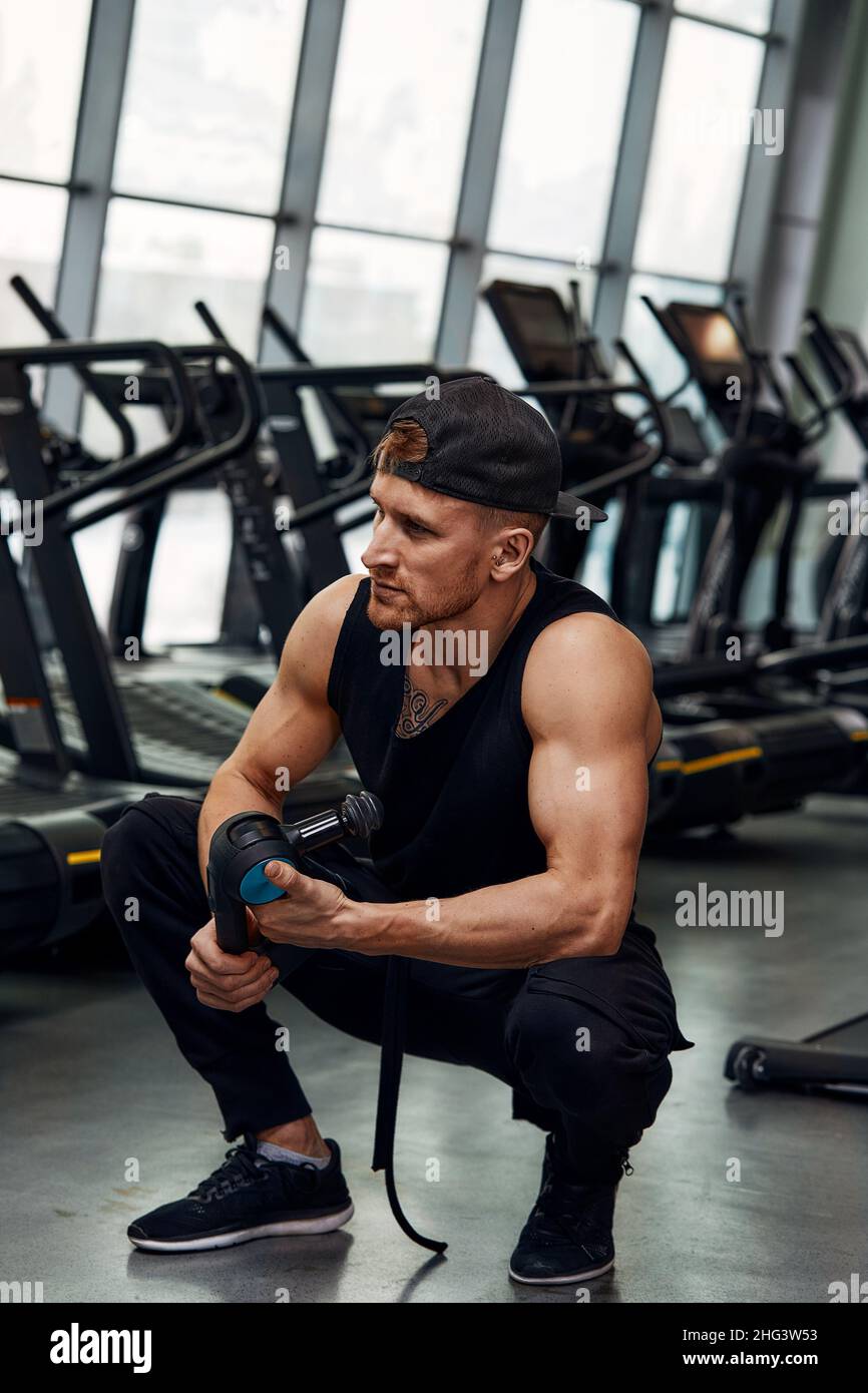 Ein junger Mann benutzt und genießt die Schlagmassagepistole, um sich zu entspannen und die Schmerzen im Körper und in den Muskeln im Fitnessstudio zu lindern. Stockfoto