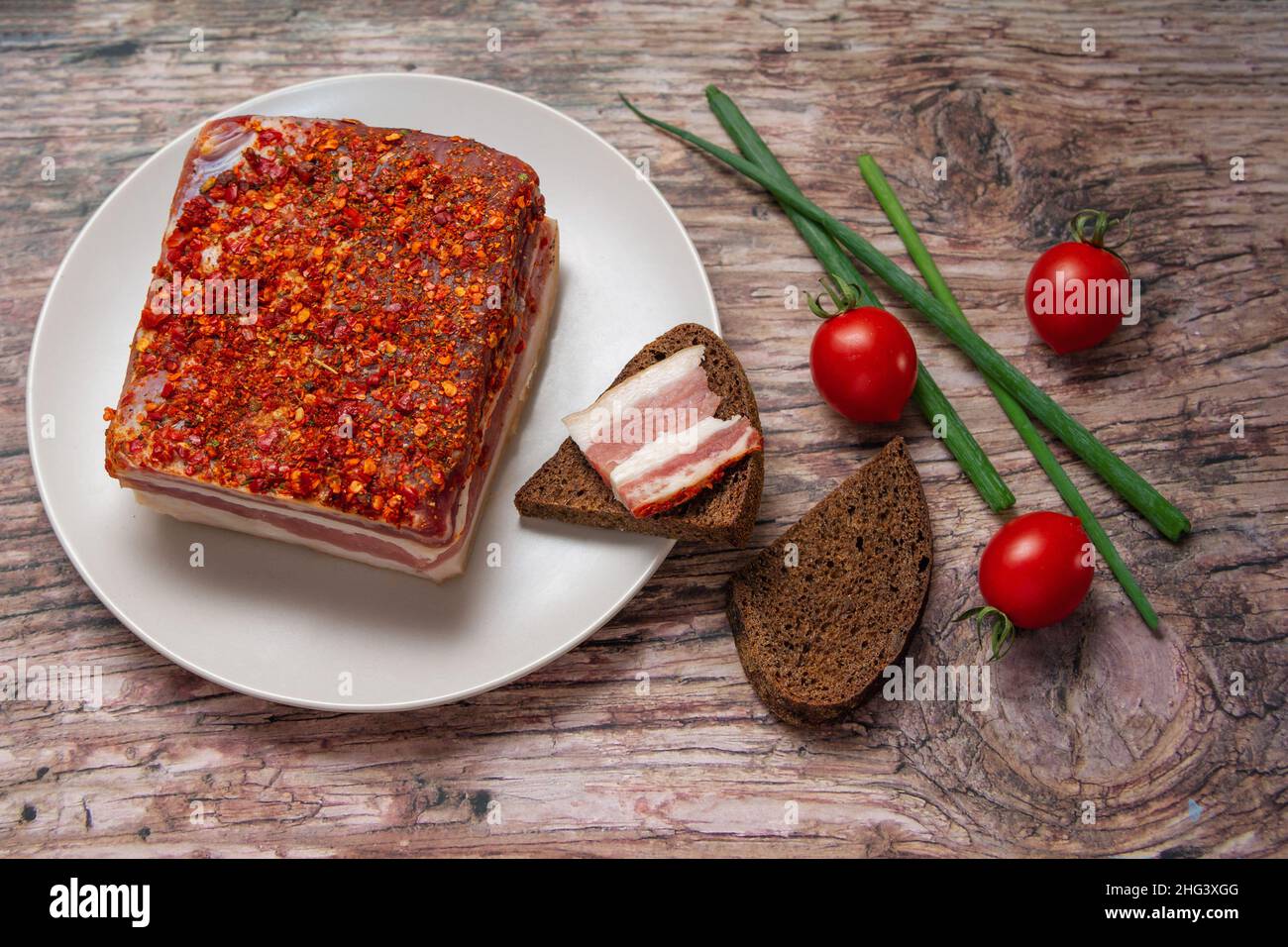 Geräucherter, würziger Speck auf einem Teller. Scheibe geräuchertes Schweinefleisch auf Brot, Kirschtomaten und grüne Zwiebel auf Holztisch. Stockfoto