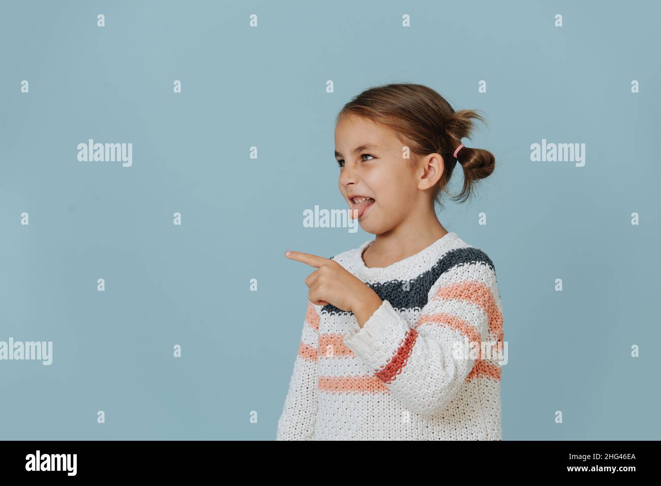 Neckende böse kleine Mädchen in gestreiften Pullover zeigt ihren Finger und streckt Zunge über blauen Hintergrund. Seitenansicht. Stockfoto