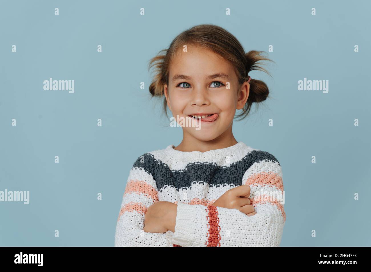 Frech kleines Mädchen in gestreiftem Pullover mit gekreuzten Händen, die Zunge über blauem Hintergrund herausragt. Stockfoto