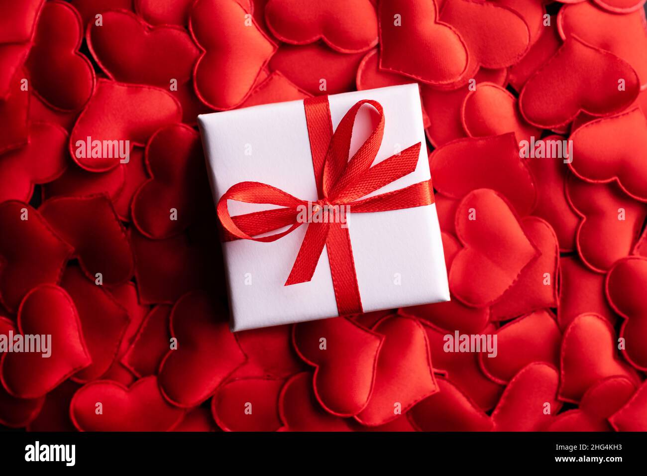 Kreatives Postkarten-Design am Valentinstag mit weißer Geschenkbox mit Schleife auf rotem Textilherzen-Hintergrund. Valentinstag und Liebeskonzept Stockfoto