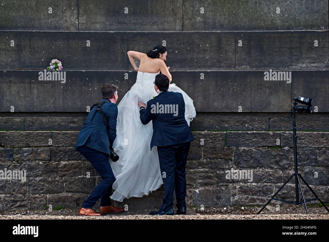 Bräutigam und Hochzeitsfotograf geben der Braut eine helfende Hand auf dem National Monument auf Calton Hill, Edinburgh, Schottland, Großbritannien. Stockfoto
