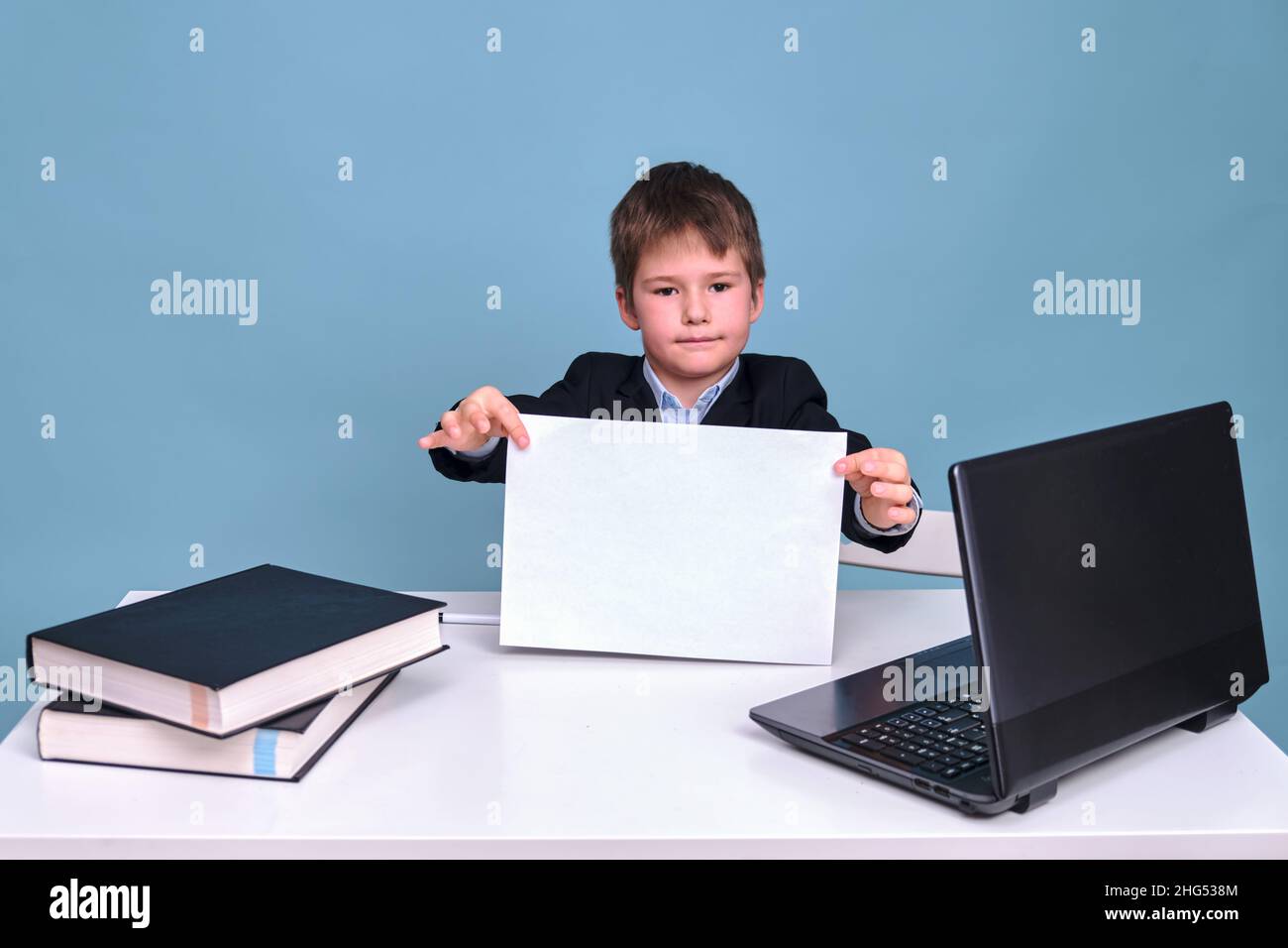 Ein Junge in einem Schulanzug mit einem Blatt A4 Papier an einem Computer während des Fernlernens, kopieren Sie Platz auf einem blauen Studiohintergrund Stockfoto