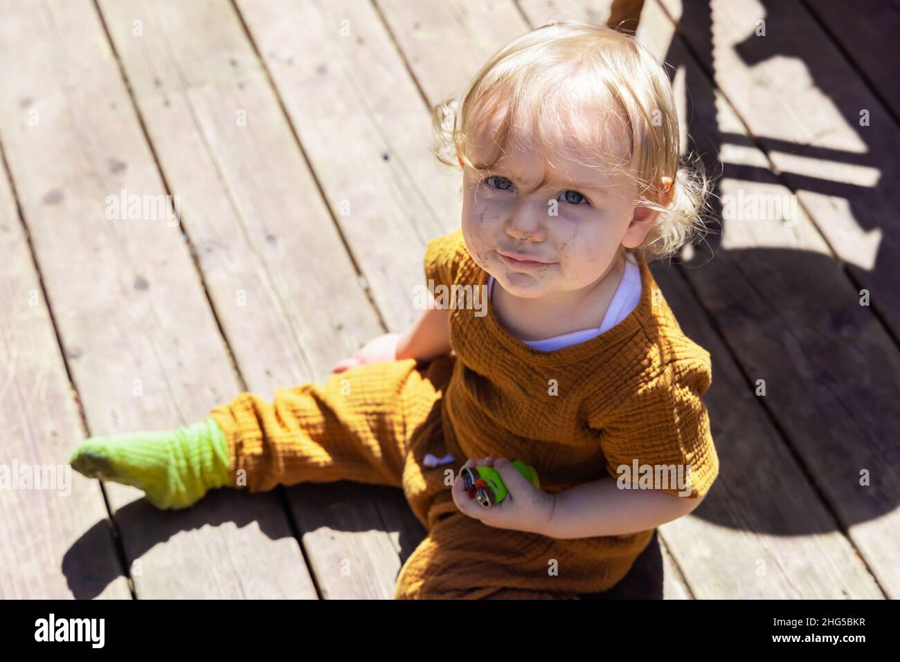 Blick aus der Perspektive eines dreijährigen, schelmischen Kleinkindes, das auf einer Holzterrasse sitzt und Feuerzeuge mit einem schmutzigen Gesicht hält, nachdem es im Hinterhof gespielt hat. Stockfoto