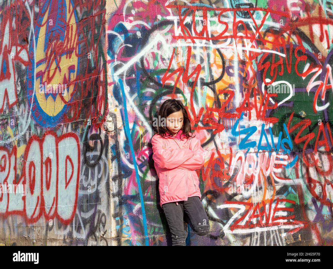 Junges Mädchen mit langen dunklen Haaren in einer rosa Jacke, die vor einer Graffiti-Wand steht. Stockfoto