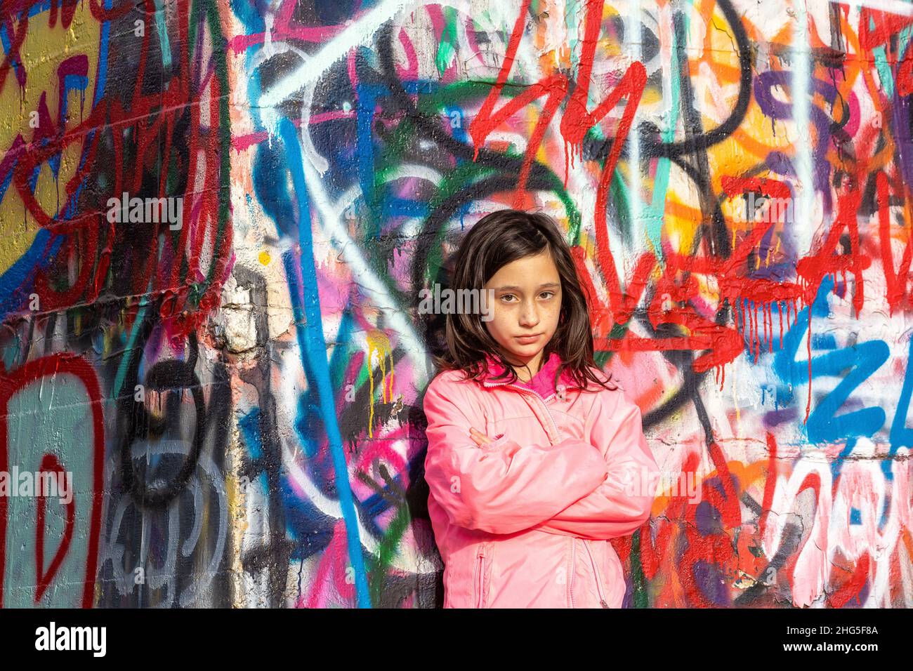 Junges Mädchen mit langen dunklen Haaren in einer rosa Jacke, die vor einer Graffiti-Wand steht. Stockfoto