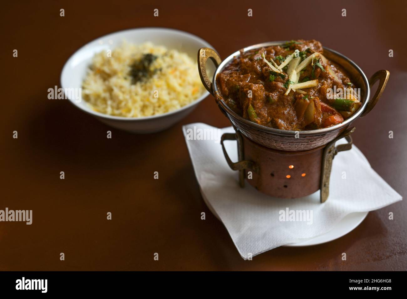Indisches Gericht mit Fleisch, Gemüse und Ingwer, serviert auf einem kleinen Metallherd mit einer Kerze wie einem Teehümer und einer Schüssel Reis auf einem braunen Tisch, Stockfoto