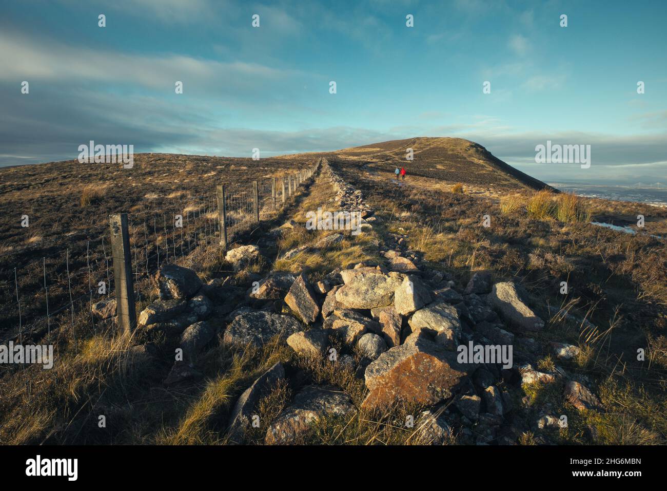 Wandern im Pentland Hills Regional Park. Im Vordergrund stehen Steine und ein Zaun, der sich in die Ferne zieht. Im Hintergrund wandern Menschen von hinten in den Hügeln. Schottland Stockfoto