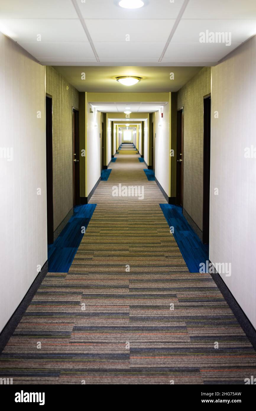 Das moderne Inn-Hotel Motel verfügt über einen leeren langen Korridor mit Teppichboden und Lampen, die eine vertikale Innenansicht der Gebäudearchitektur beleuchten Stockfoto