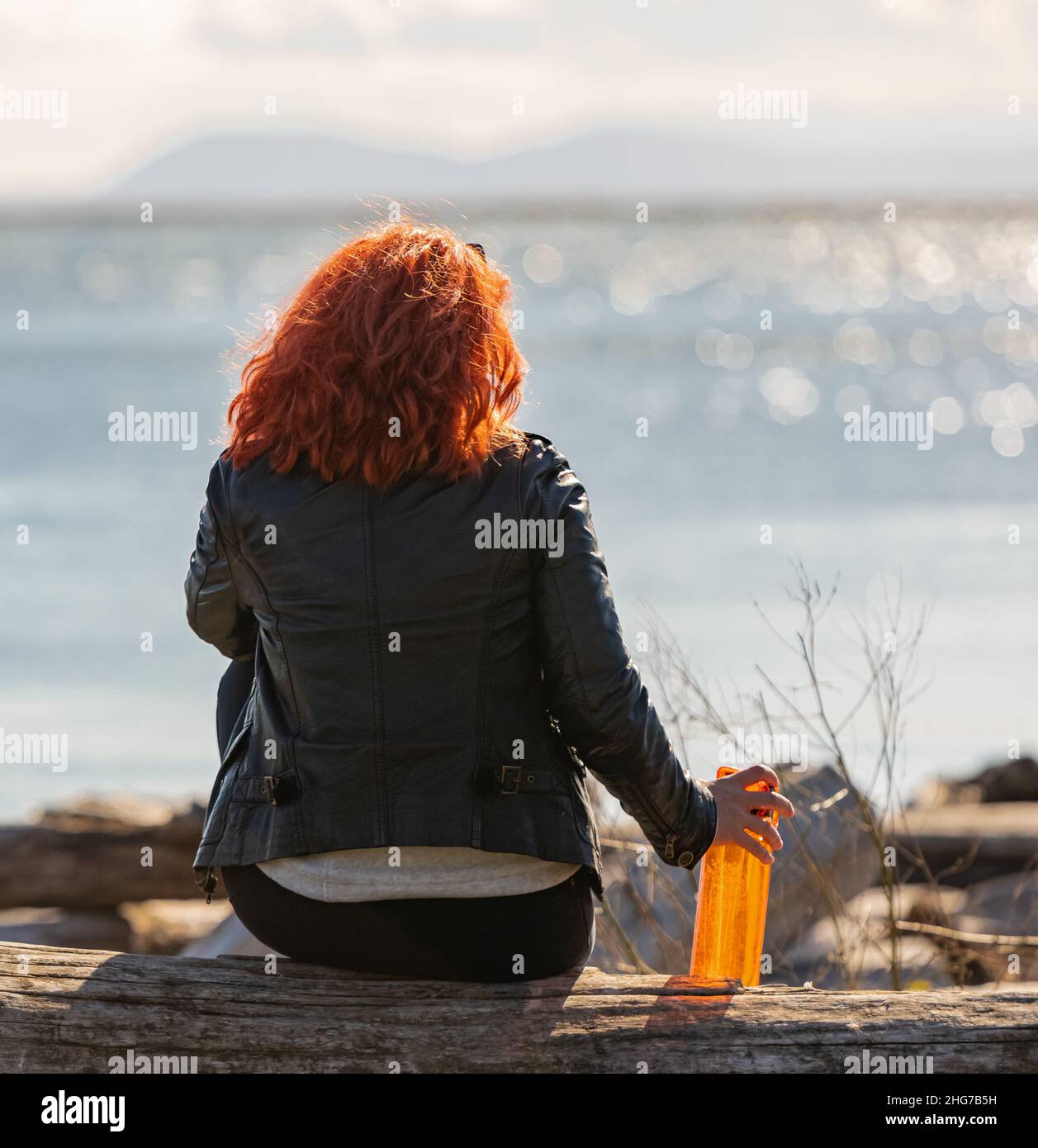 Rückansicht einer jungen Frau, die am Strand sitzt und das Meer mit einer Wasserflasche in der Hand betrachtet. Meditation in der Natur. Blick auf die Straße, Reisefoto, Stockfoto