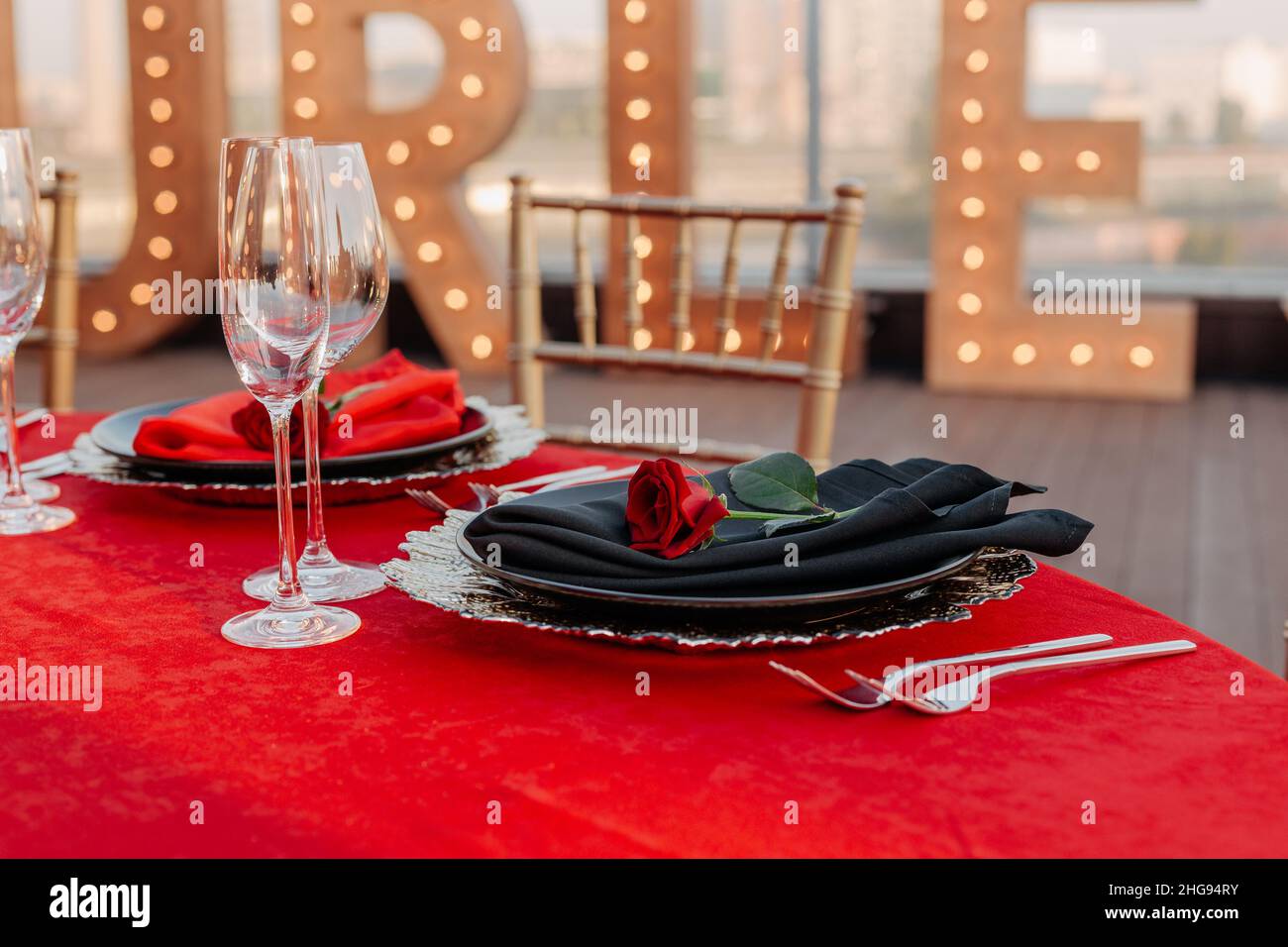 Gäste können an einem Tisch in Schwarz, Rot und Gold Bankett machen. Elegantes Abendessen: Dekor, Tischdecke, Teller mit Servietten und frischen Rosen, Gläser, Besteck. Motto-Party-Feier auf dem Dach, im Freien Stockfoto