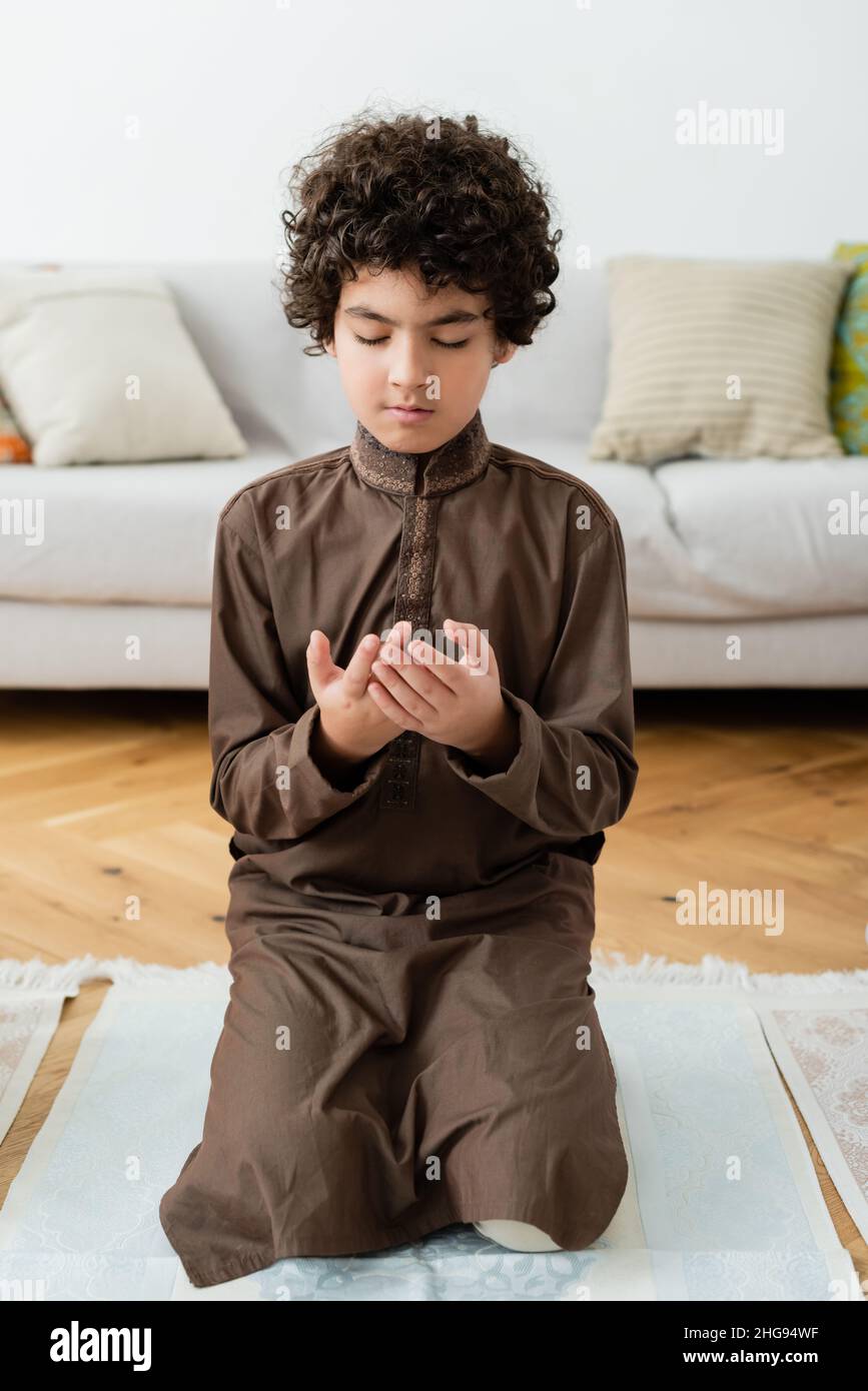 Lockiges arabisches Kind, das zu Hause auf einem traditionellen Teppich betet Stockfoto