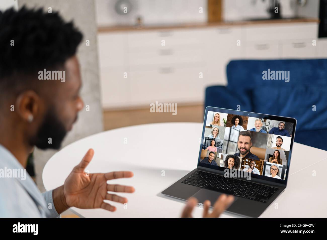 Remote-Meeting per Videoanruf. Multirassischer Mann, der in der Küche sitzt und die App auf dem Laptop für die Online-Kommunikation mit einer Gruppe verschiedener multirassischer Menschen verwendet. Rückansicht Stockfoto