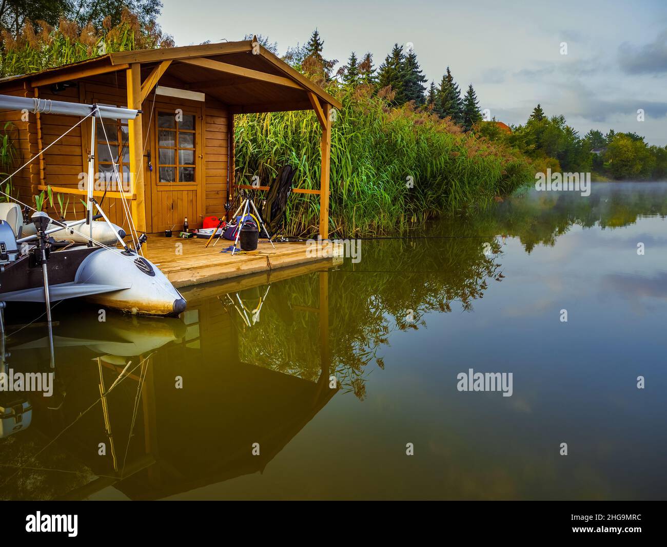 Ein Fischerhaus und ein Schlauchboot auf dem Fluss, versteckt in einem Schilf. Kleines Holzhaus zum Angeln, entspannen und ruhige Zeit, reflektiert in einem Fluss Wasser in Stockfoto