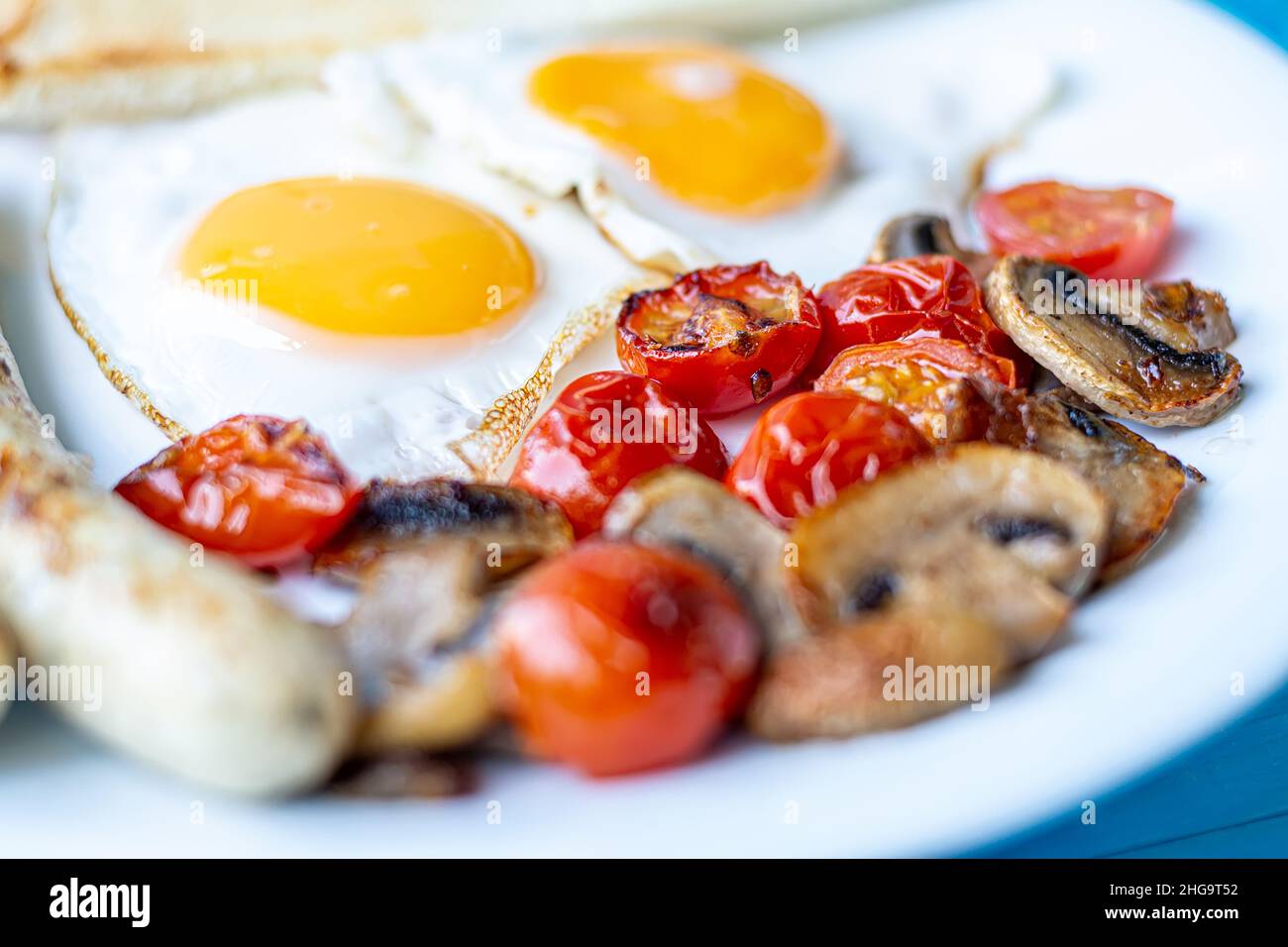 Frühstück mit Spiegeleiern, gegrillten Würstchen, gebratenen Pilzen und Kirschtomaten, Makroaufnahme auf einem weißen Teller. Stockfoto