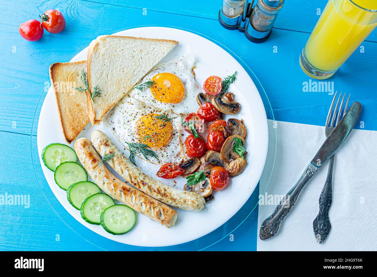 Frühstück mit Spiegeleiern, gegrillten Würstchen, gebratenen Pilzen und Kirschtomaten, frischen Gurkenscheiben und knusprigem Toast auf einem weißen Teller auf einem hölzernen BL Stockfoto