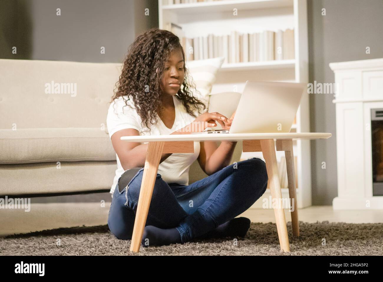 Hübsche junge Frau mit gefalteten Beinen sitzt auf dem Teppich und tippt auf ihrem Computer. Die junge Frau schaut sich die Filmliste auf ihrem Laptop an. Nahaufnahme. Startseite Hintergrund . Hochwertige Fotos Stockfoto