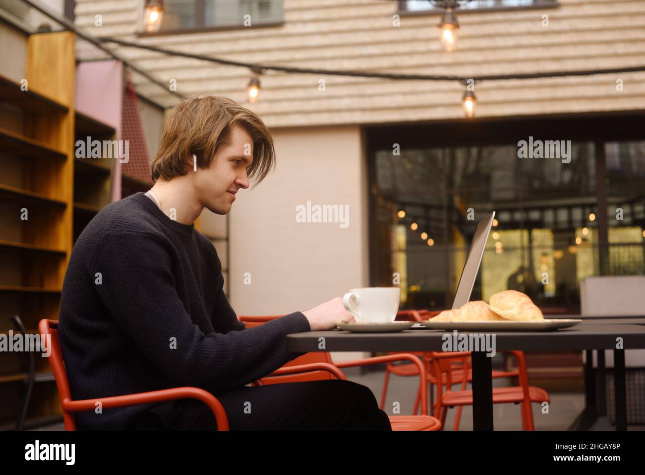 Mann trinkt eine Tasse Kaffee in einem Restaurant auf der Terrasse. Freiberufler im Freien mit einem Laptop, hat Frühstück mit Croissants. Businessman Looks and drea Stockfoto