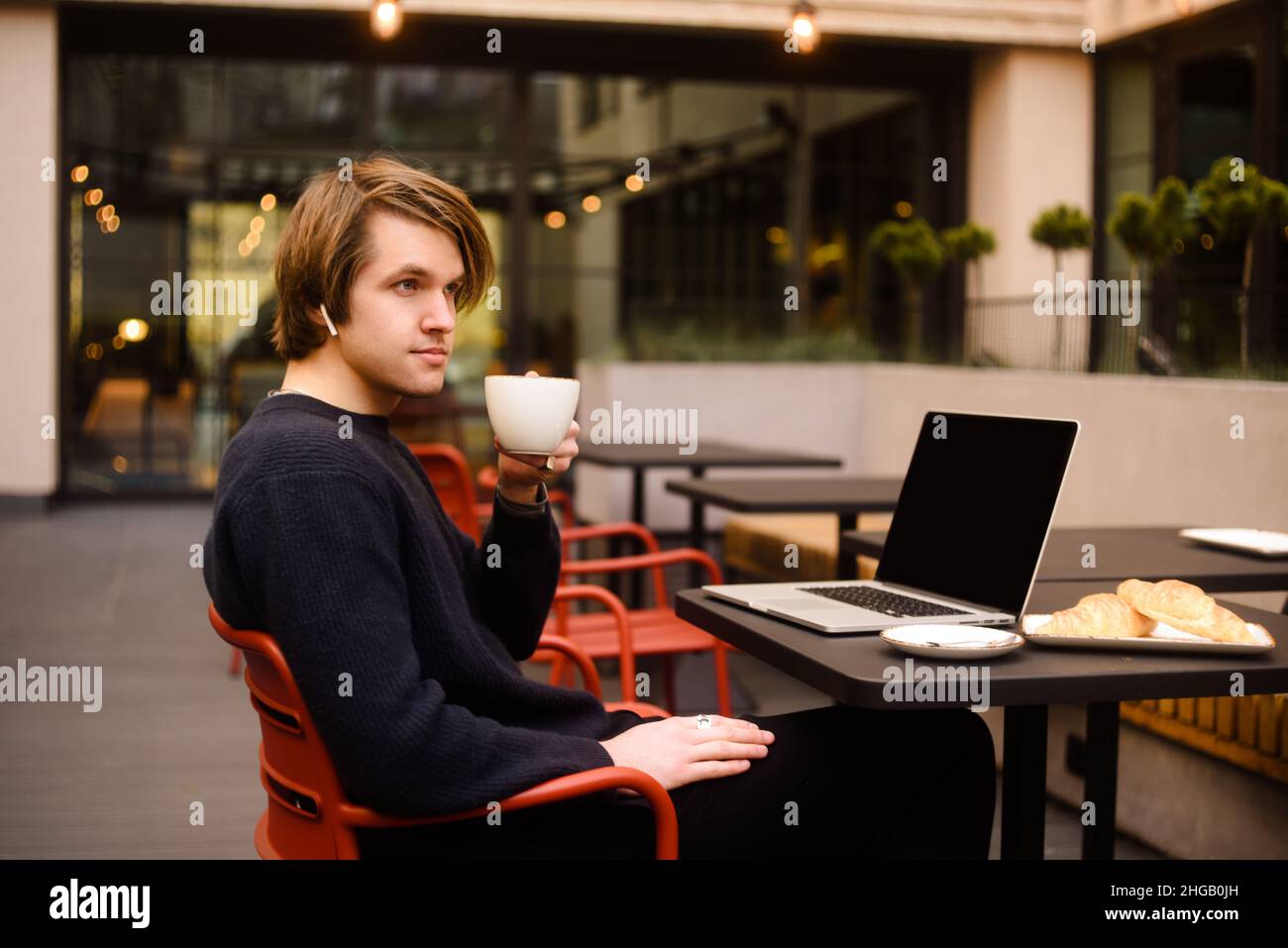 Mann trinkt eine Tasse Kaffee in einem Restaurant auf der Terrasse. Freiberufler im Freien mit einem Laptop, hat Frühstück mit Croissants. Businessman Looks and drea Stockfoto