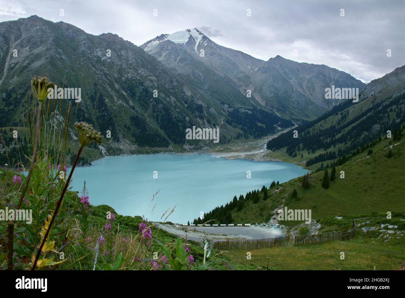 Großer Almaty See in Bergen mit Straße und Bergrücken und Gipfeln im Hintergrund und Blumen im Vordergrund Stockfoto