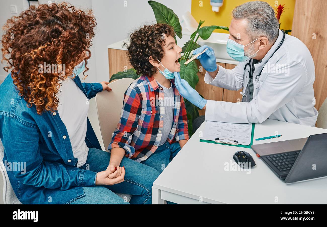 Kind mit ihrem Elternteil bei Kinderarzttermin. Der Hausarzt untersucht mit dem Inspektionsspatel den Hals des Jungen Stockfoto