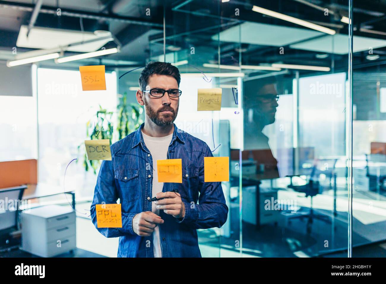 Fokussierter und denkender Geschäftsmann, der auf bunten Notizen an einer Glaswand schreibt, erstellt der Unternehmer eine visuelle To-Do-Liste, Geschäftsziele für Stockfoto