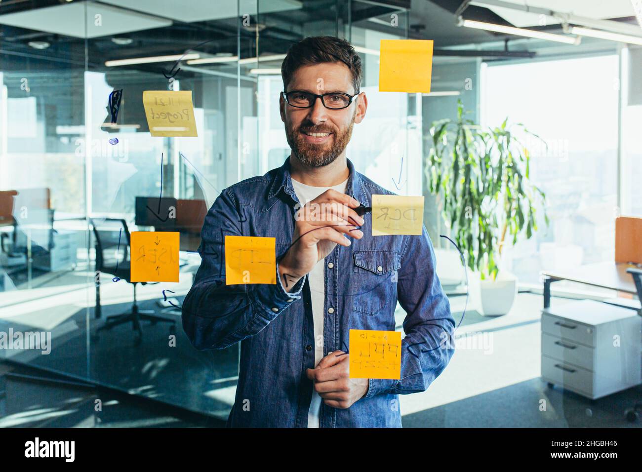Fokussierter und denkender Geschäftsmann, der auf bunten Notizen an einer Glaswand schreibt, erstellt der Unternehmer eine visuelle To-Do-Liste, Geschäftsziele für Stockfoto