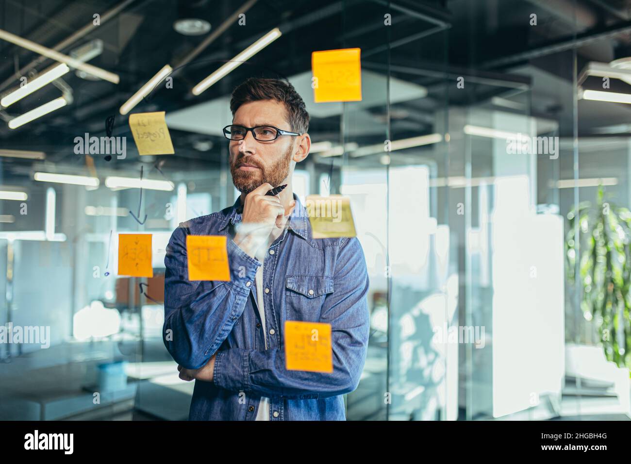 Fokussierter und denkender Geschäftsmann, der auf bunten Notizen an einer Glaswand schreibt, erstellt der Unternehmer eine visuelle To-Do-Liste, Geschäftsziele für Stockfoto