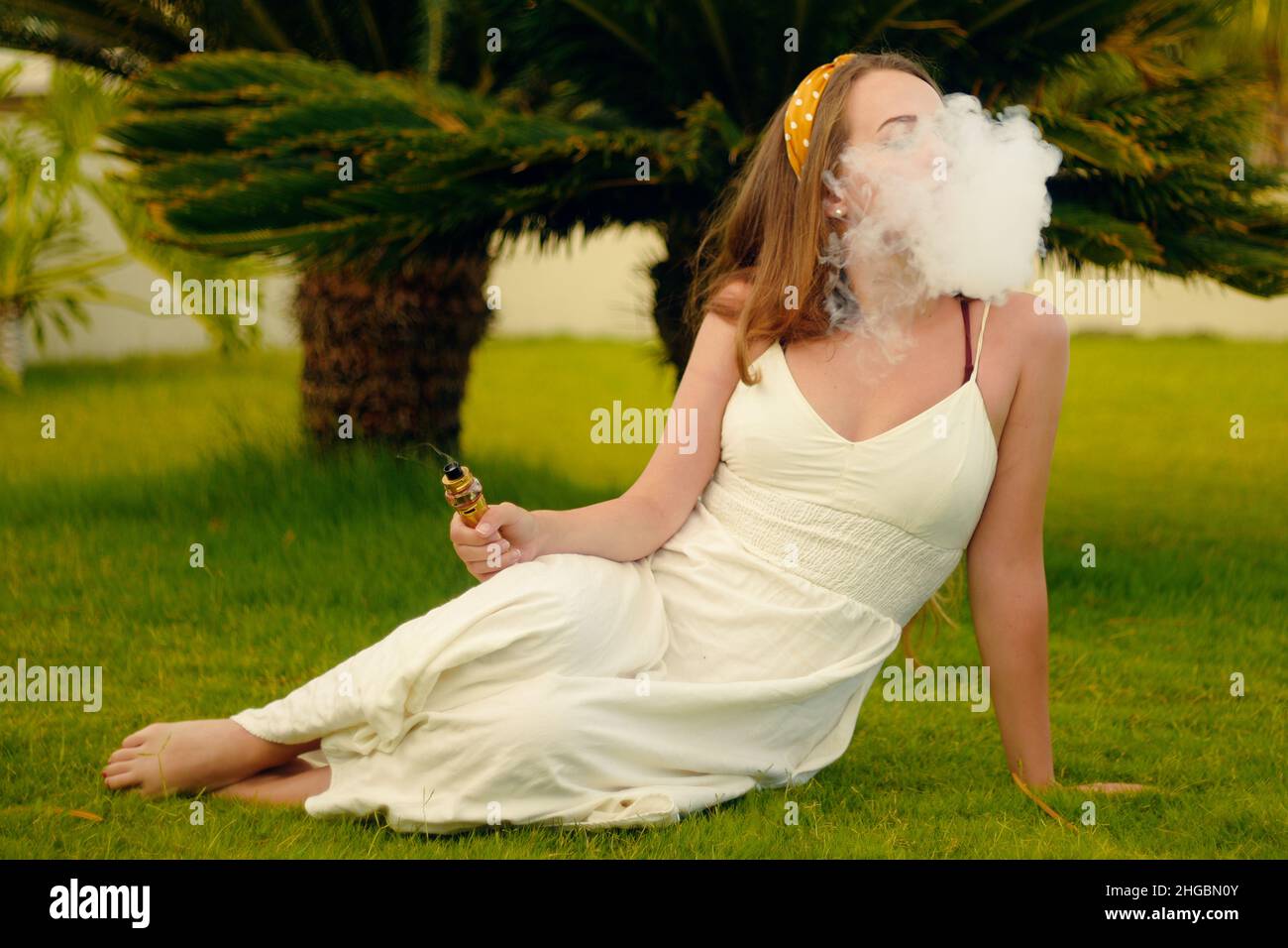 Eine Vape-Frau im Urlaub, unter einer Palme. Ruhe, Rauchen, Nikotin, Genuss, Gefahr. Flüssigkeit abdampfen. Dampfende THC e Zigarette. Stockfoto