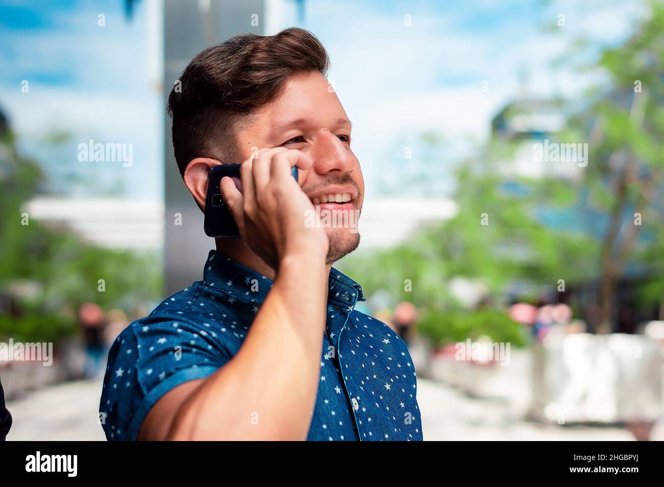 Der junge Mann, hispanic Latino, mit Bart und kurzen Haaren, gekleidet in ein blaues Kurzarmhemd, lächelt und redet am Telefon, mit dem Himmel und sma Stockfoto
