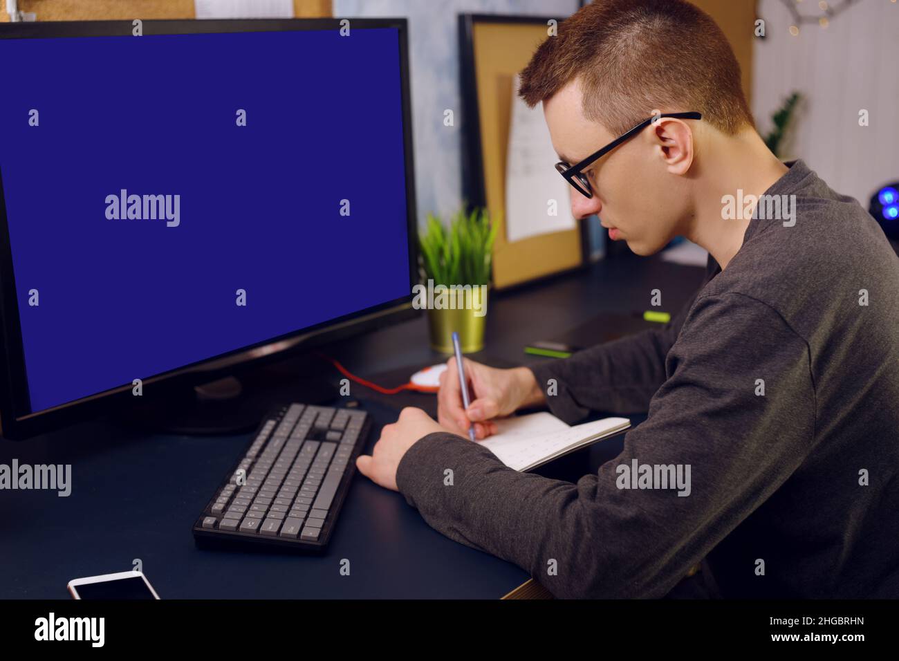 Ein Mann arbeitet zu Hause, schreibt mit einem Stift auf ein Notizbuch. Ein Mann schaut auf den grünen Bildschirm des Computers, Hausaufgaben. Ein Mann arbeitet als Freiberufler, lernt neue te Stockfoto
