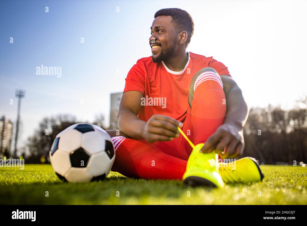 Fröhlicher multikultureller hispanischer Fußballspieler, der lächelt und an einem sonnigen Tag einen roten, sportlichen Anzug trägt Stockfoto