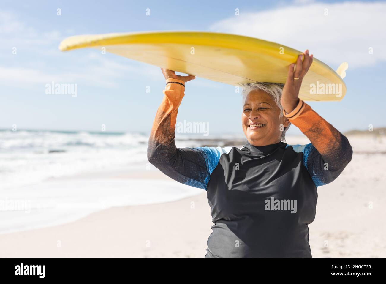 Glückliche ältere Frau aus dem Birazial genießt den Ruhestand, während sie am sonnigen Strand Surfbrett auf dem Kopf trägt Stockfoto