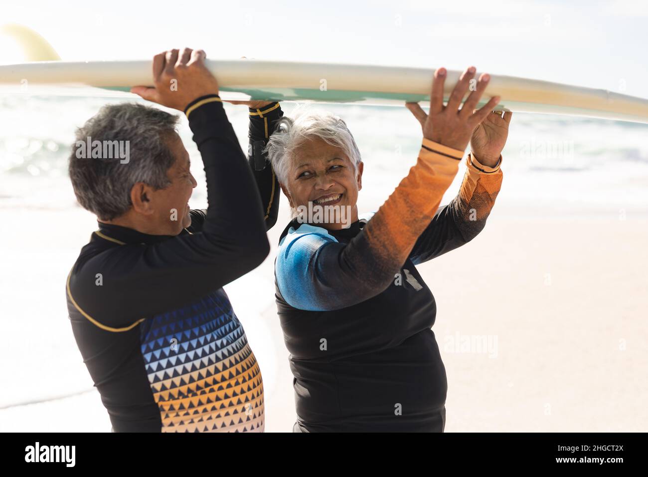 Seitenansicht eines glücklichen, mehrrassigen älteren Paares, das am sonnigen Strand Surfbrett über die Köpfe trug Stockfoto