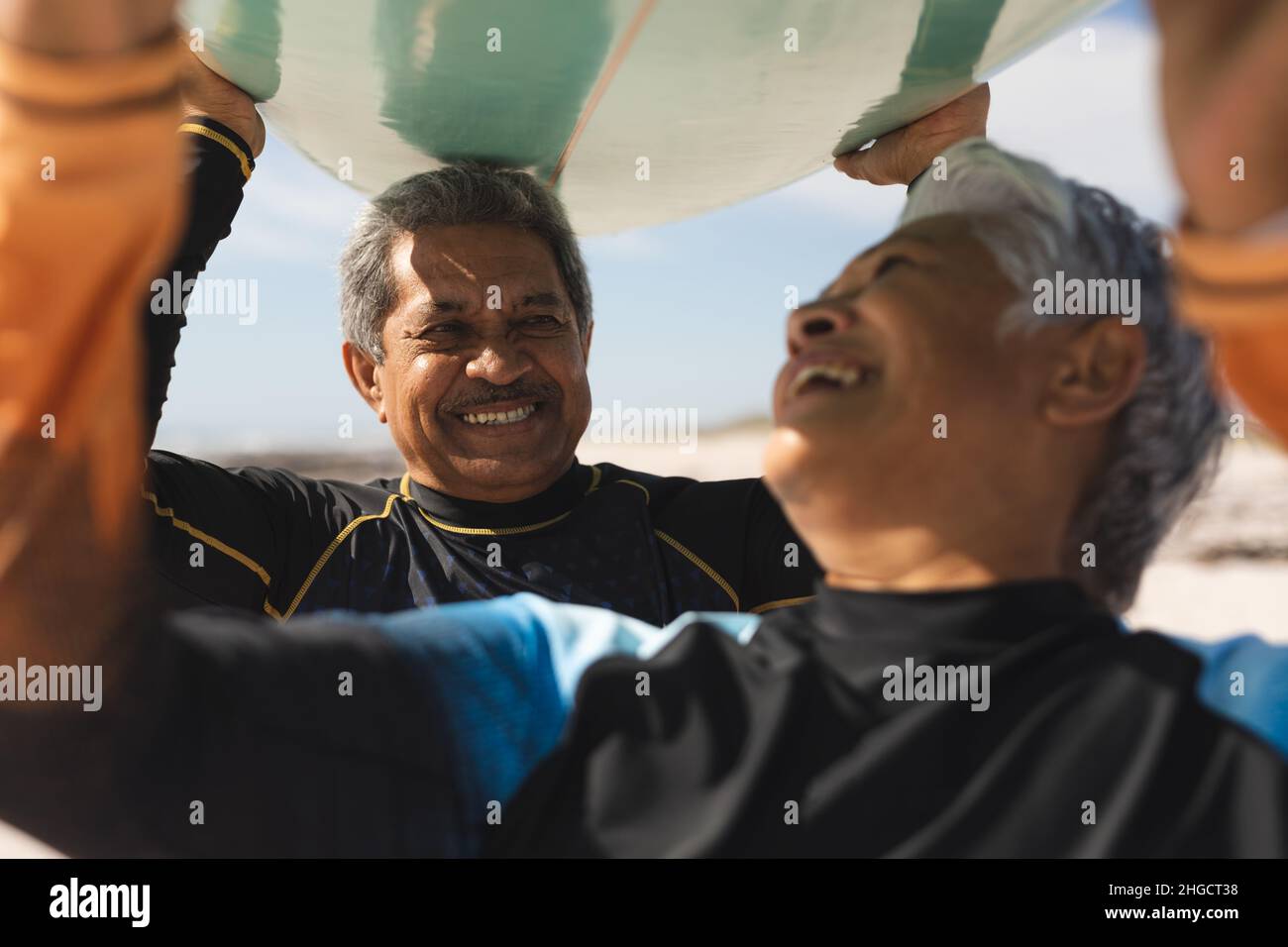Fröhliches, mehrrassiges Senioren-Paar im Ruhestand, das am Strand Surfbrett über die Köpfe trägt Stockfoto