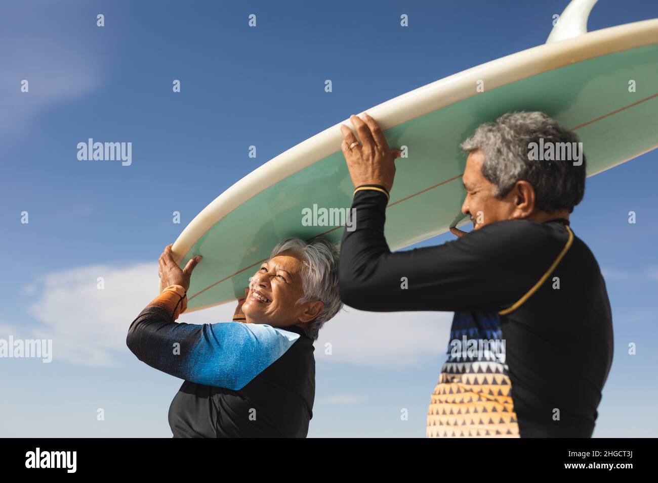 Blick aus der unteren Perspektive auf ein glückliches, multirassisches Seniorenpaar, das Surfbrett auf dem Kopf gegen den blauen Himmel trägt Stockfoto