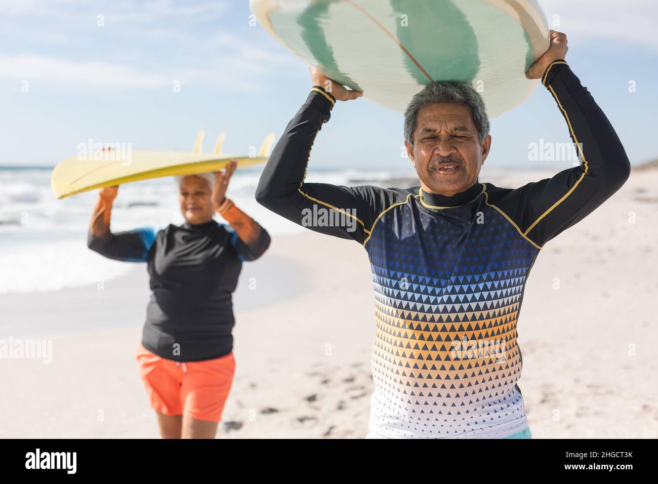 Multirassisches Seniorenpaar genießt gemeinsam den Ruhestand, während es Surfbretter über den Köpfen am Strand trägt Stockfoto