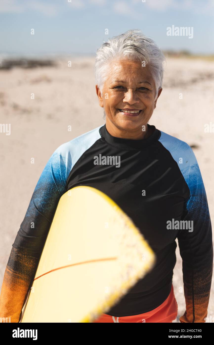 Porträt einer lächelnden älteren Birazialfrau mit kurzen weißen Haaren, die am sonnigen Strand Surfbrett hält Stockfoto