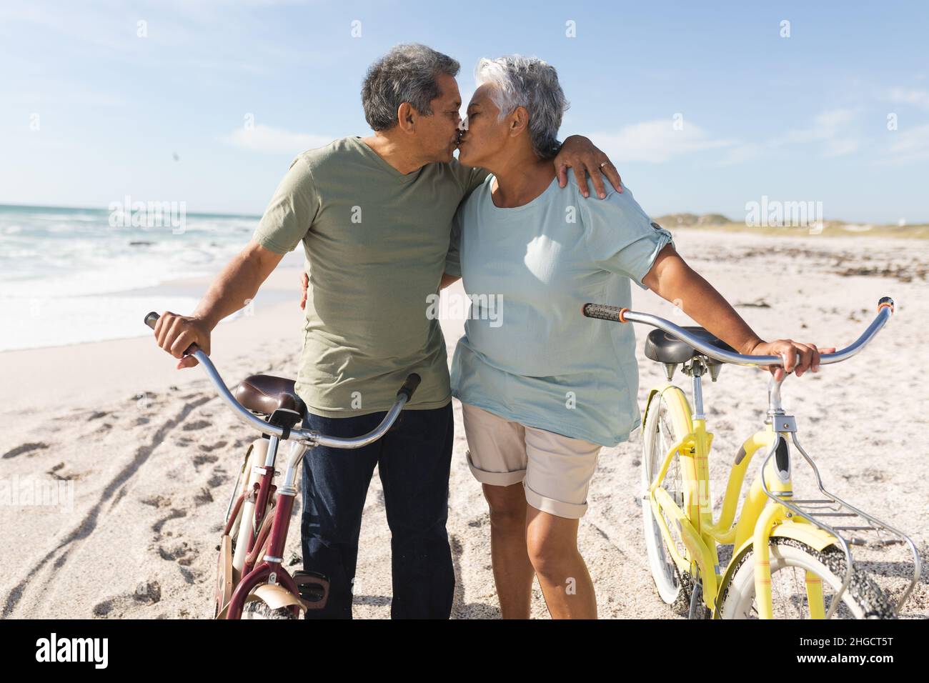 Zärtliches multirassisches Seniorenpaar, das sich küsst, während es mit Zyklen am sonnigen Strand gegen den Himmel steht Stockfoto