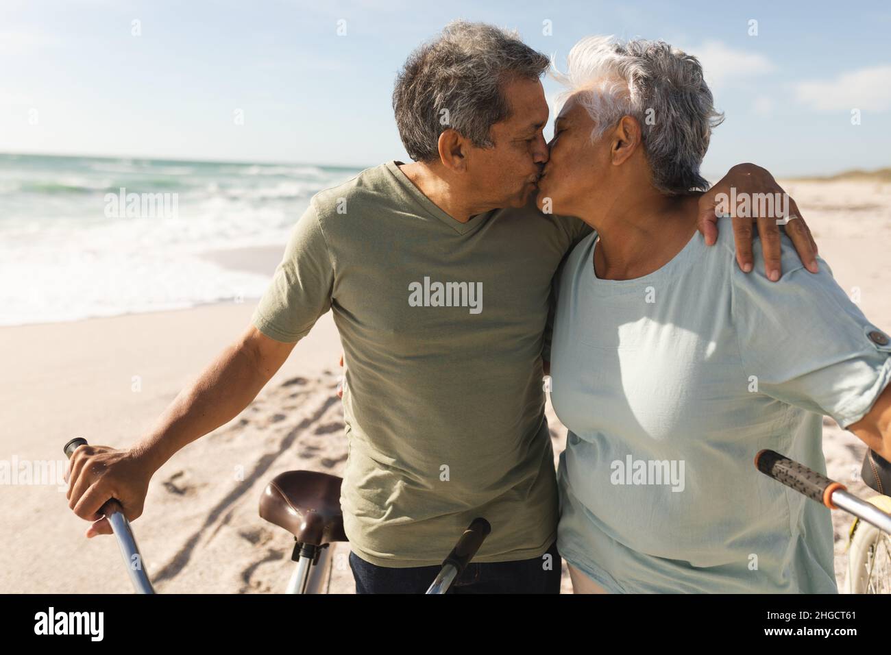 Rentnerpaar mit mehreren Rassen, das sich während eines sonnigen Tages mit Fahrrädern am Strand küsst Stockfoto